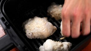 A hand places coated chicken pieces into an air fryer basket