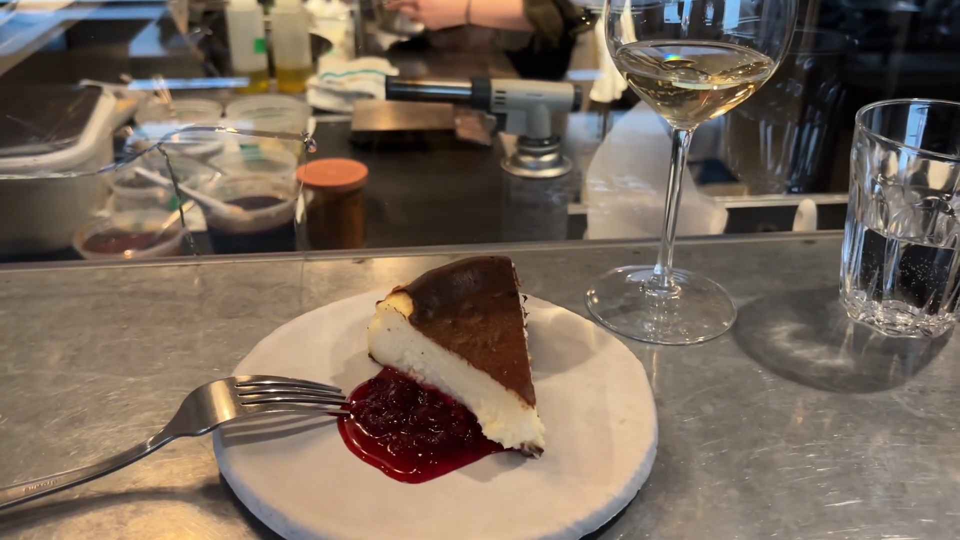 Slice of Basque cheesecake with cherry sauce on a plate, next to a glass of white wine at a restaurant counter