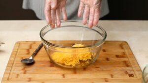 Shredded cheddar cheese is being added to a glass mixing bowl on a wooden cutting board