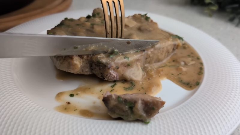 A fork and knife are cutting into a pork chop covered in a thick maple Dijon glaze on a white plate