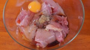 Raw chicken thighs in a glass bowl are topped with soy sauce, egg, spices, and seasonings, ready to be mixed for marinating