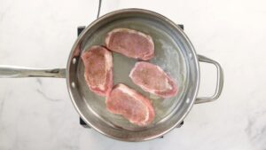 Four seasoned pork chops are being seared in a stainless steel pan with oil