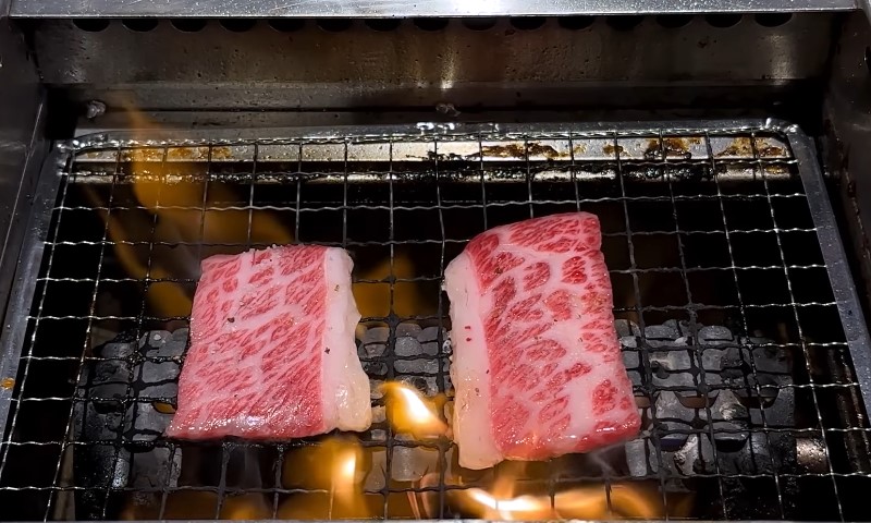 Two pieces of meat sizzling on a grill, surrounded by smoke and flames