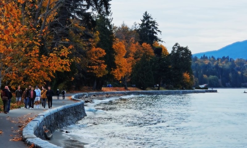 People walk along a lakeside path on an overcast day, bordered by trees with vibrant orange and green foliage