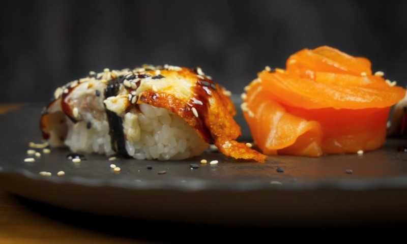 A close-up of sushi on a dark plate, featuring a nigiri with grilled fish, nori, and sesame seeds