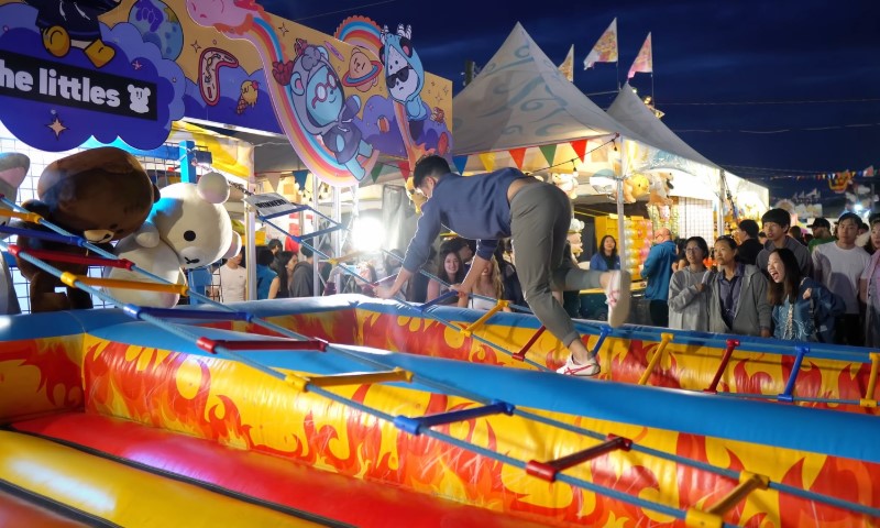 Man in casual clothes balances on an inflatable ladder in a vibrant, illuminated carnival setting
