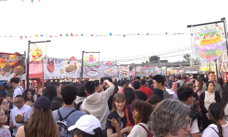 A bustling outdoor market scene with a large, diverse crowd. Colorful banners and food stalls line the path