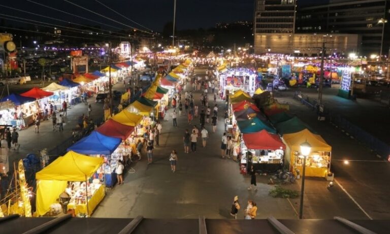 Night market scene with colorful tents lining both sides of a wide pathway crowded with people