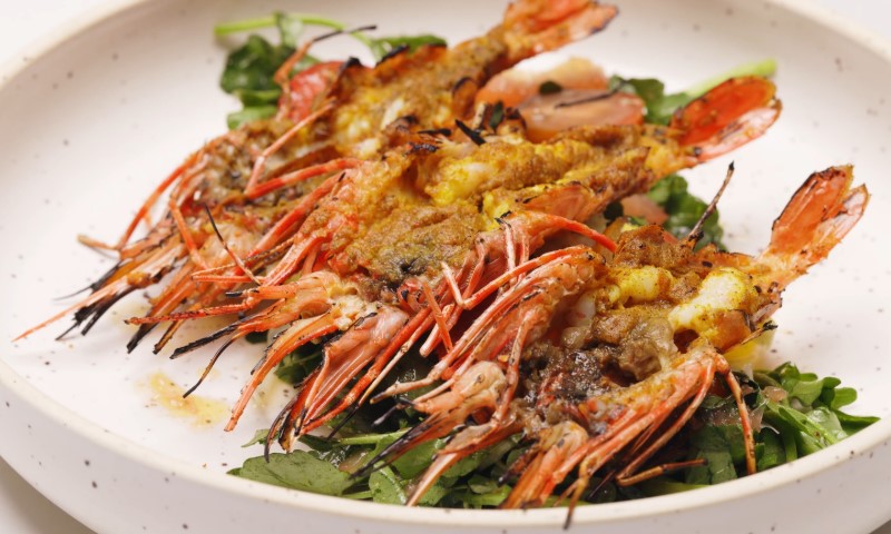 Grilled shrimp with spicy marinade, displayed on a white plate, are set on a bed of fresh greens