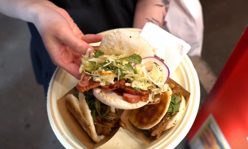 A person holds a plate with three bao buns filled with vibrant toppings like cabbage, herbs, and meat