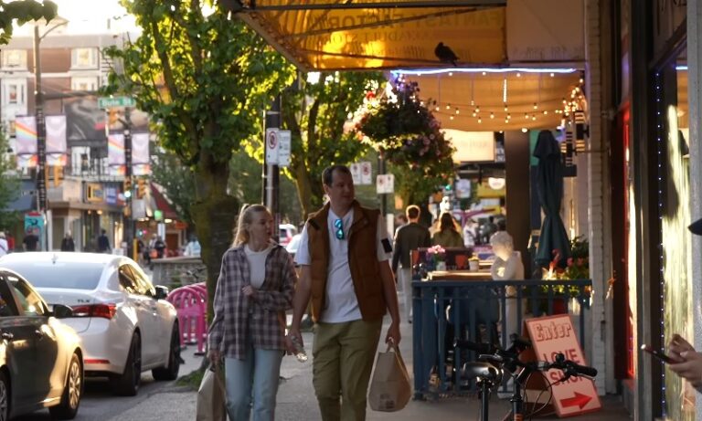 A couple walks along a lively urban street at dusk, holding shopping bags
